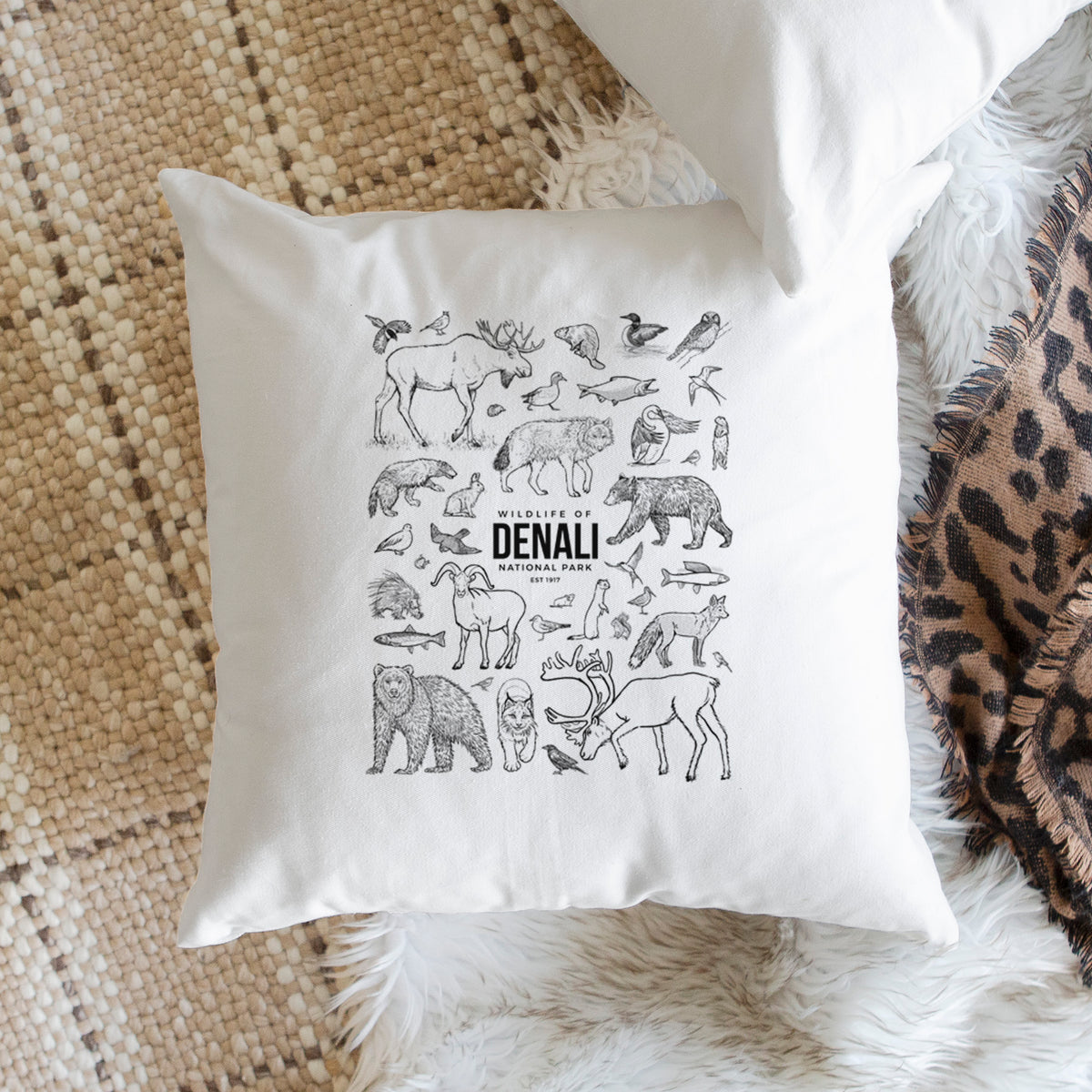White pillow cover printed with a collection of black ink wildlife illustrations surrounding the words Denali National Park, on a woven chair.