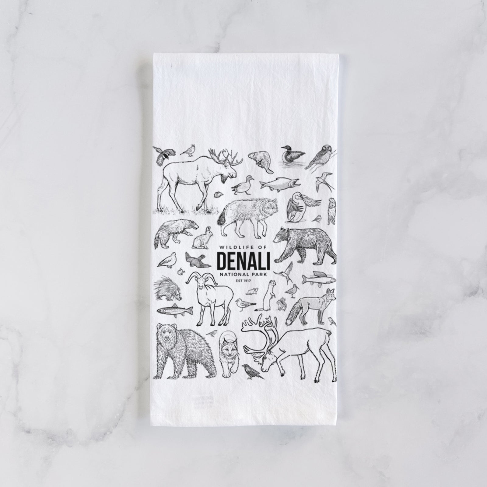 White cotton tea towel printed with a collection of black ink wildlife illustrations surrounding the words Denali National Park, on a marble counter beside a round wooden cutting board and greenery.