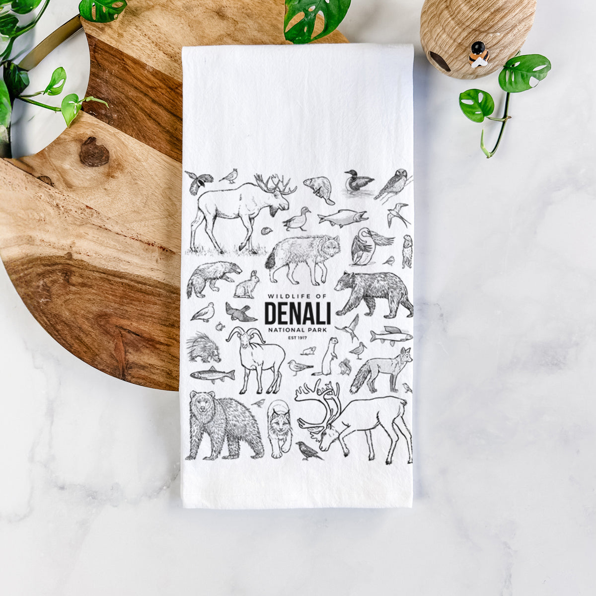 White cotton tea towel printed with a collection of black ink wildlife illustrations surrounding the words Denali National Park, on a marble counter beside a round wooden cutting board and greenery.