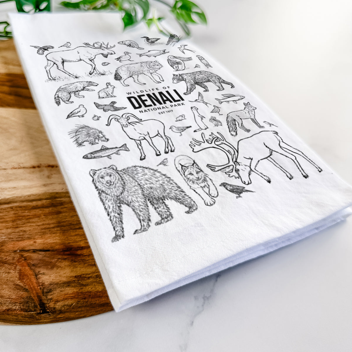 Close-up of a folded white tea towel on a wooden board, showing a collection of black ink wildlife illustrations surrounding the words Denali National Park.