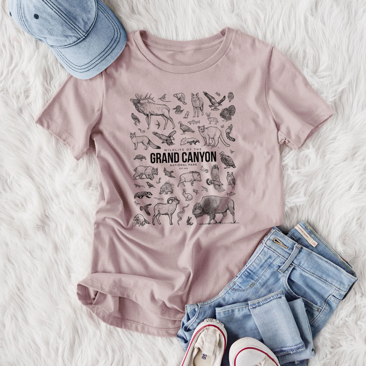 Women's relaxed fit cotton t-shirt printed with a collection of black ink wildlife illustrations surrounding the words Grand Canyon National Park, styled flat on a white faux fur rug with jeans.