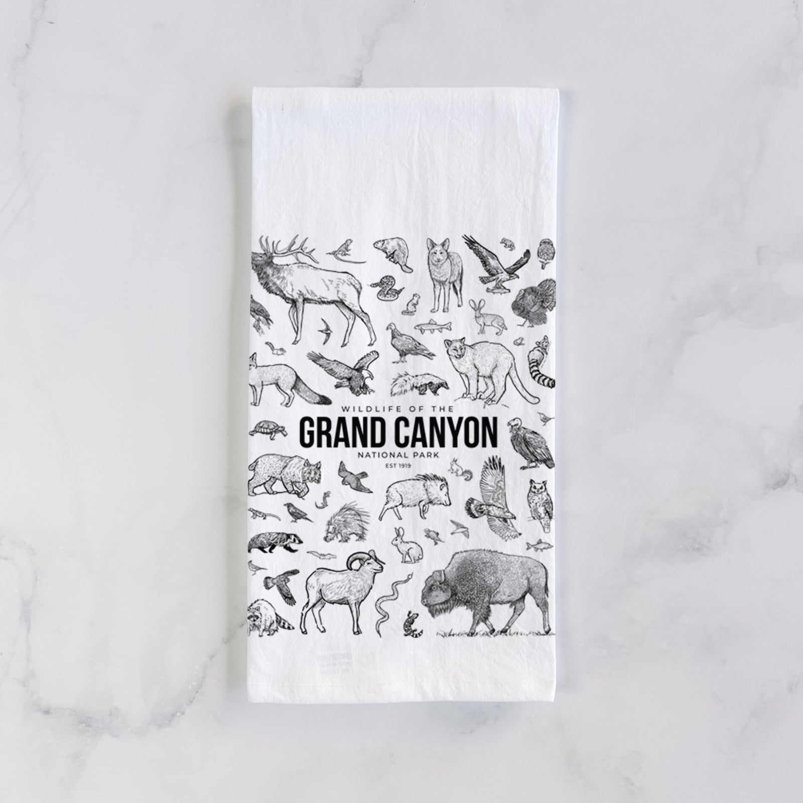 White cotton tea towel printed with a collection of black ink wildlife illustrations surrounding the words Grand Canyon National Park, on a marble counter beside a round wooden cutting board and greenery.