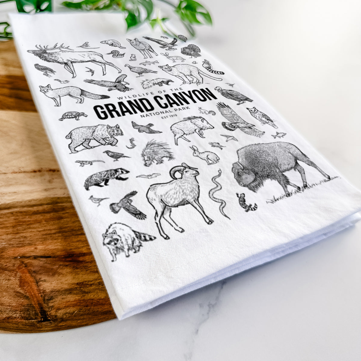 Close-up of a folded white tea towel on a wooden board, showing a collection of black ink wildlife illustrations surrounding the words Grand Canyon National Park.