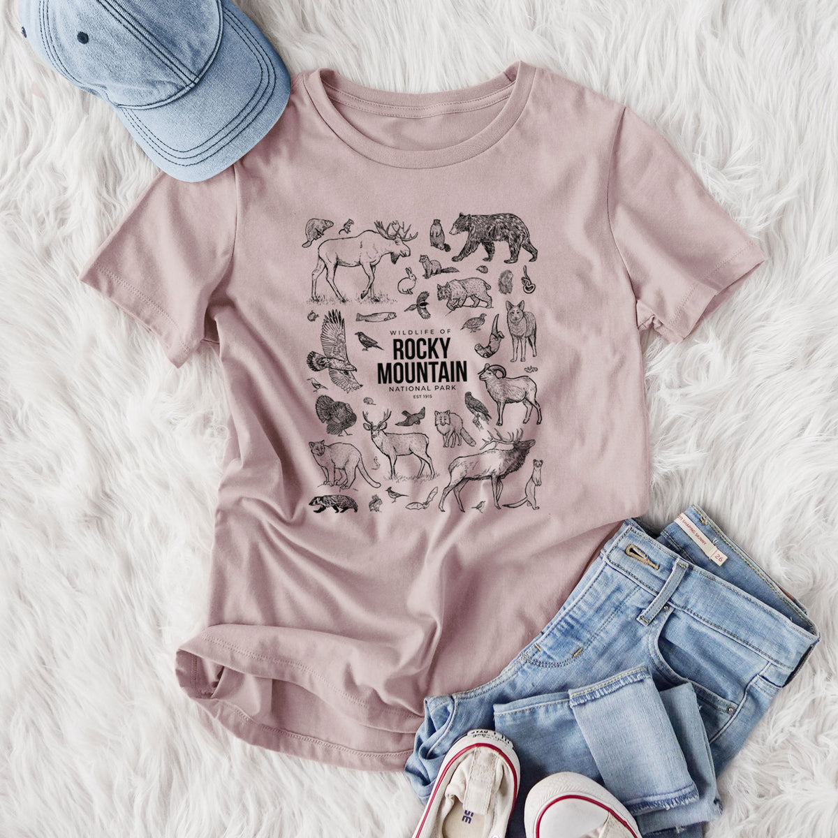 Women's relaxed fit cotton t-shirt printed with a collection of black ink wildlife illustrations surrounding the words Rocky Mountain National Park, styled flat on a white faux fur rug with jeans.