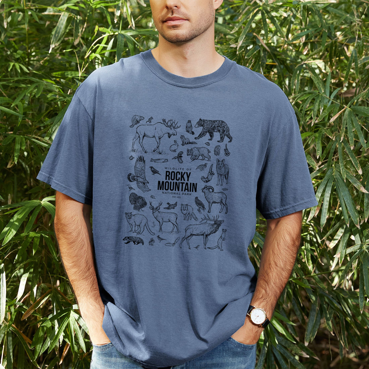 Man wearing a Comfort Colors t-shirt printed with a collection of black ink wildlife illustrations surrounding the words Rocky Mountain National Park, outdoors.