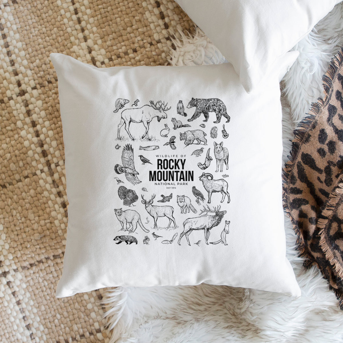 White pillow cover printed with a collection of black ink wildlife illustrations surrounding the words Rocky Mountain National Park, on a woven chair.
