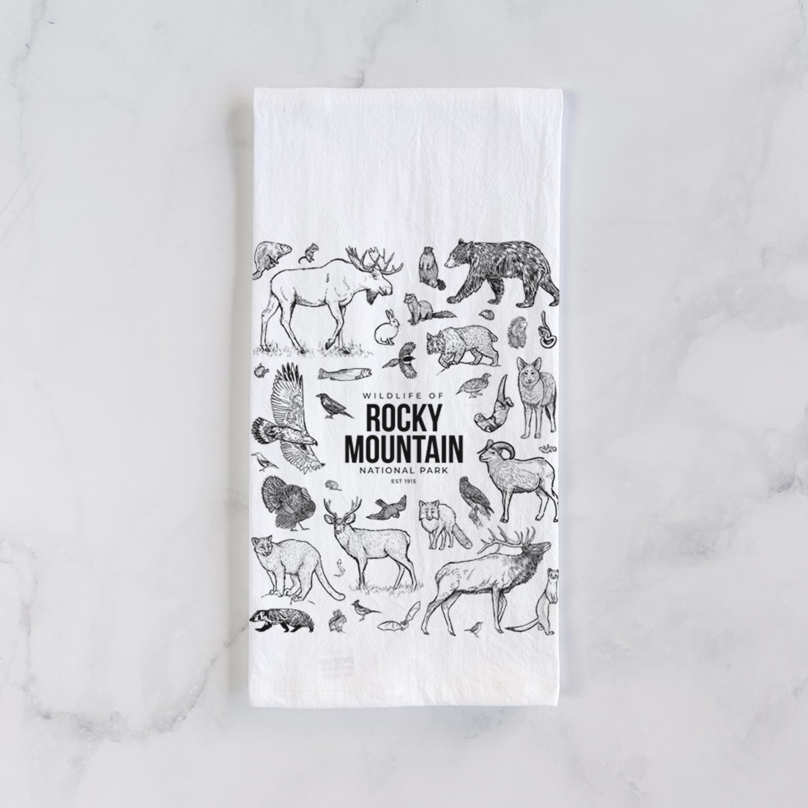 White cotton tea towel printed with a collection of black ink wildlife illustrations surrounding the words Rocky Mountain National Park, on a marble counter beside a round wooden cutting board and greenery.
