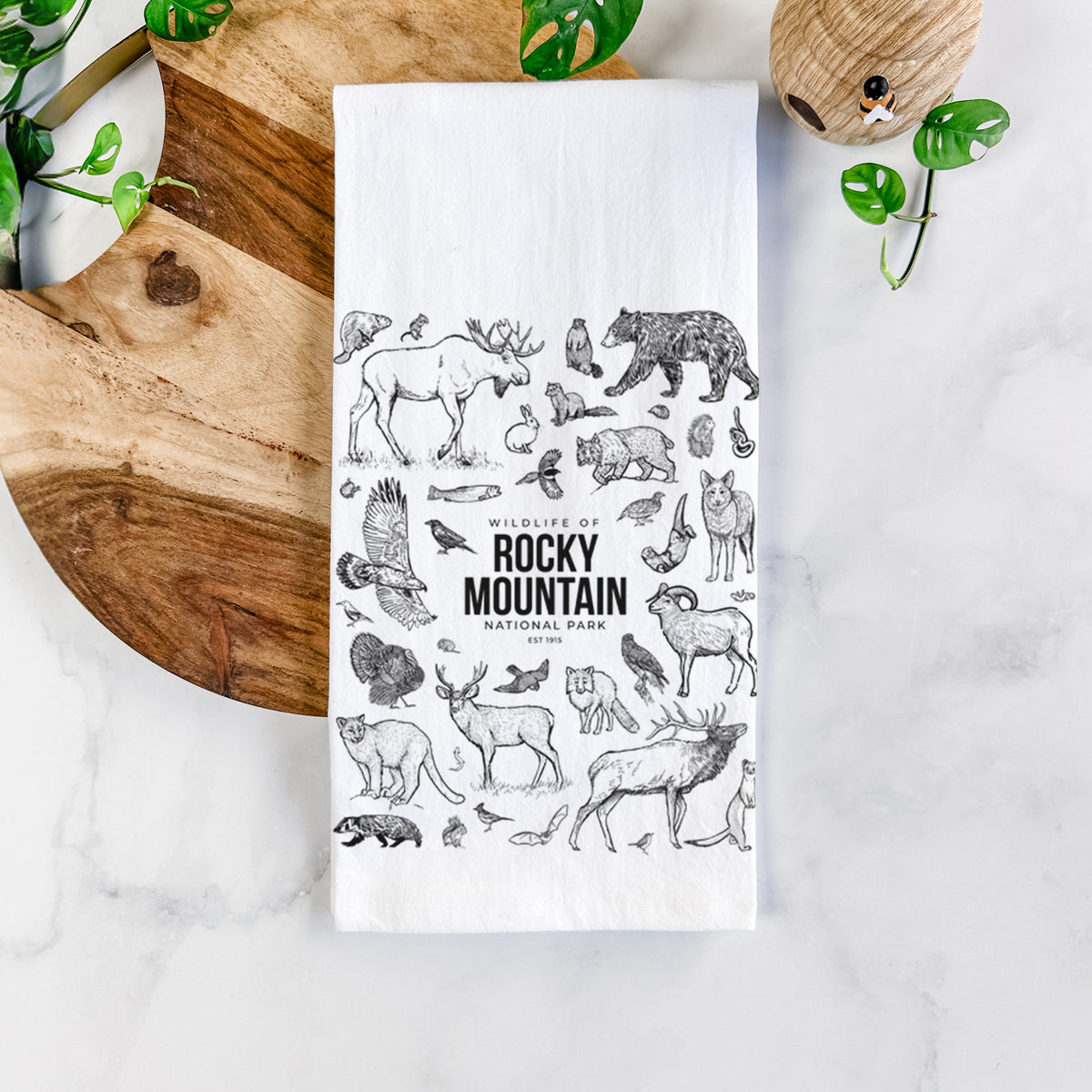 White cotton tea towel printed with a collection of black ink wildlife illustrations surrounding the words Rocky Mountain National Park, on a marble counter beside a round wooden cutting board and greenery.