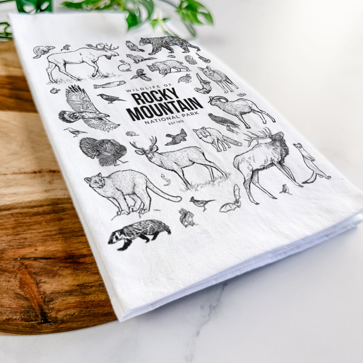 Close-up of a folded white tea towel on a wooden board, showing a collection of black ink wildlife illustrations surrounding the words Rocky Mountain National Park.