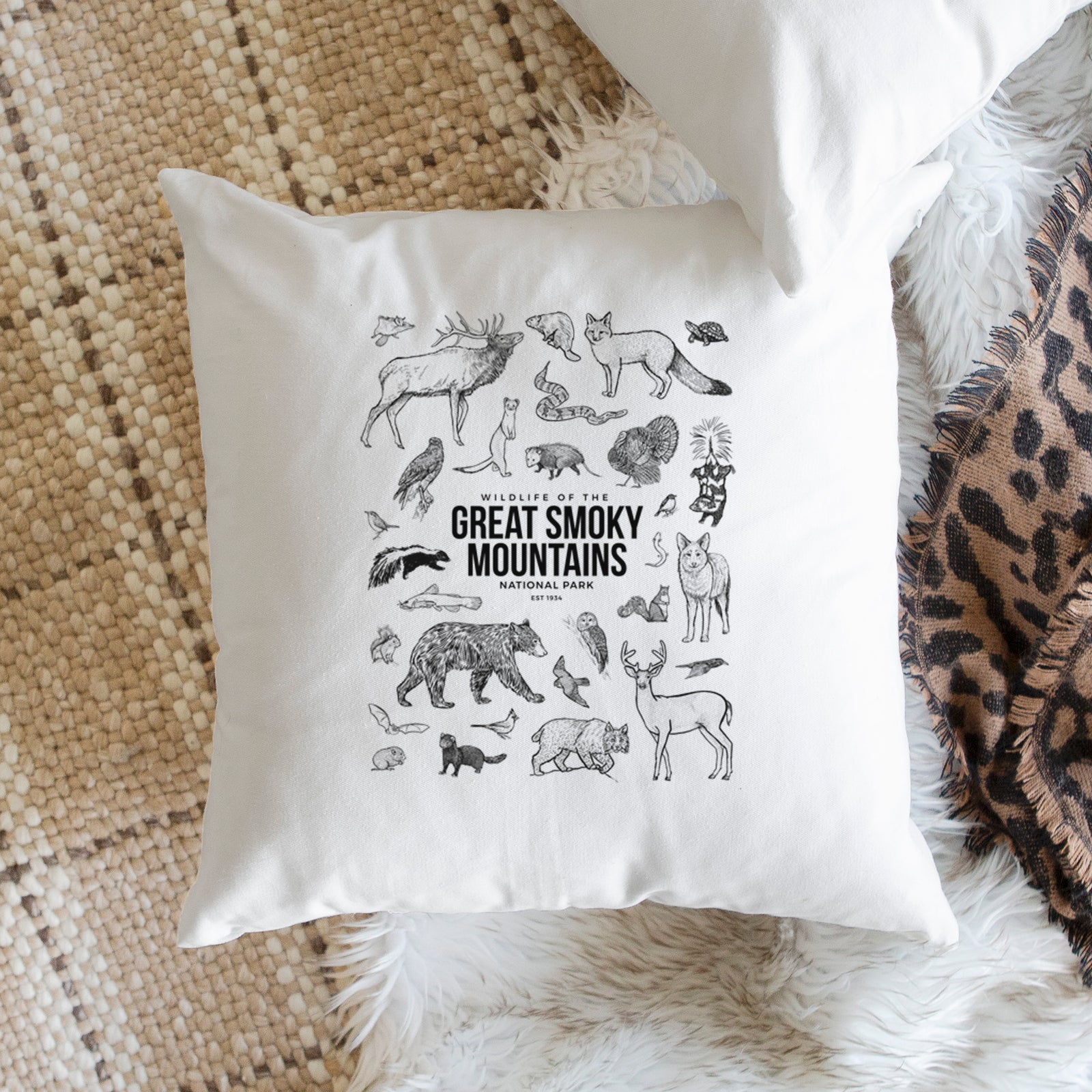 White pillow cover printed with a collection of black ink wildlife illustrations surrounding the words Great Smoky Mountains National Park, on a bench beside plants.