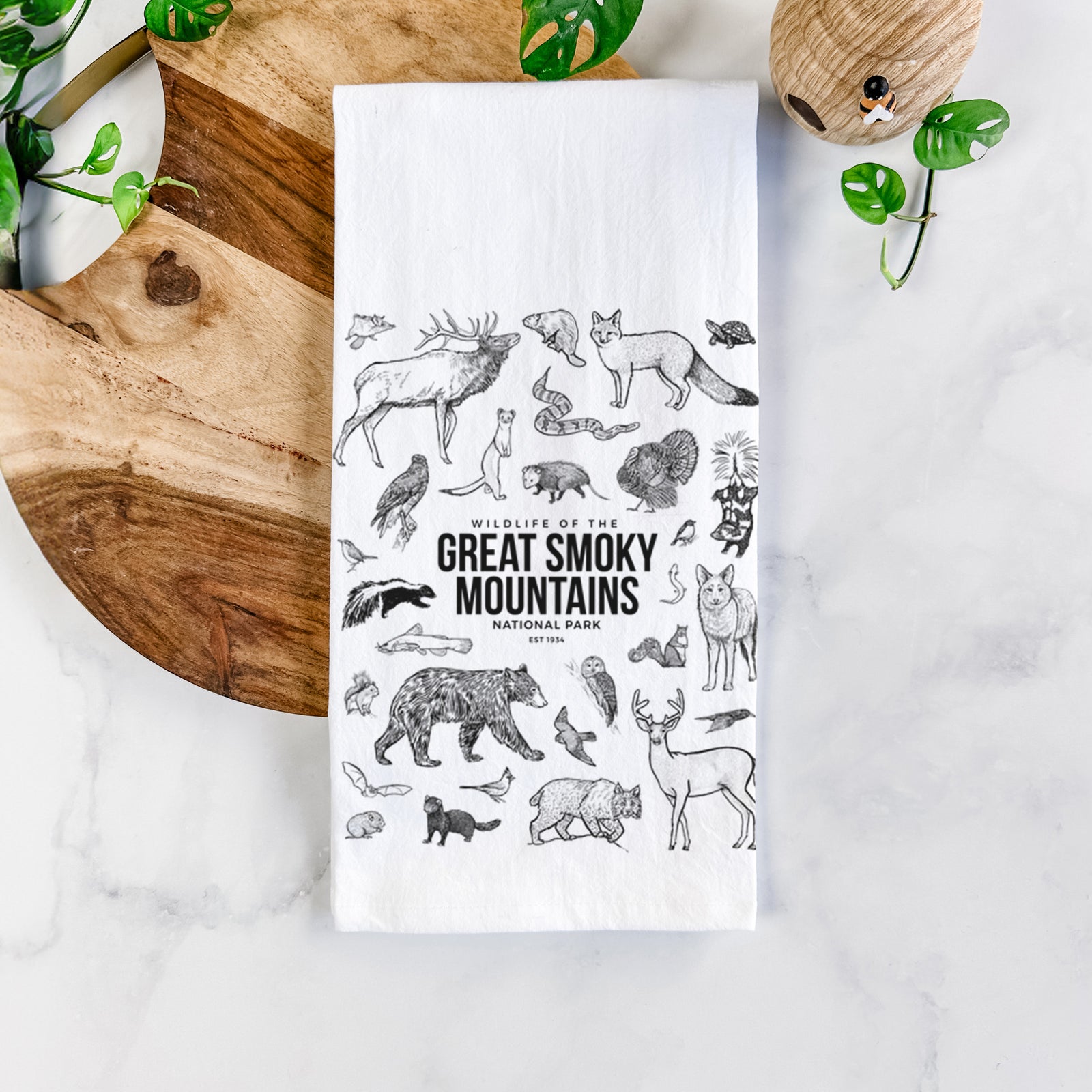 White cotton tea towel printed with a collection of black ink wildlife illustrations surrounding the words Great Smoky Mountains National Park, on a marble counter beside a round wooden cutting board and greenery.