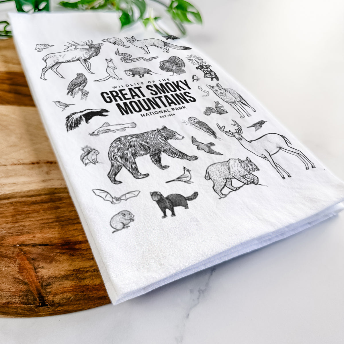 Close-up of a folded white tea towel on a wooden board, showing a collection of black ink wildlife illustrations surrounding the words Great Smoky Mountains National Park.