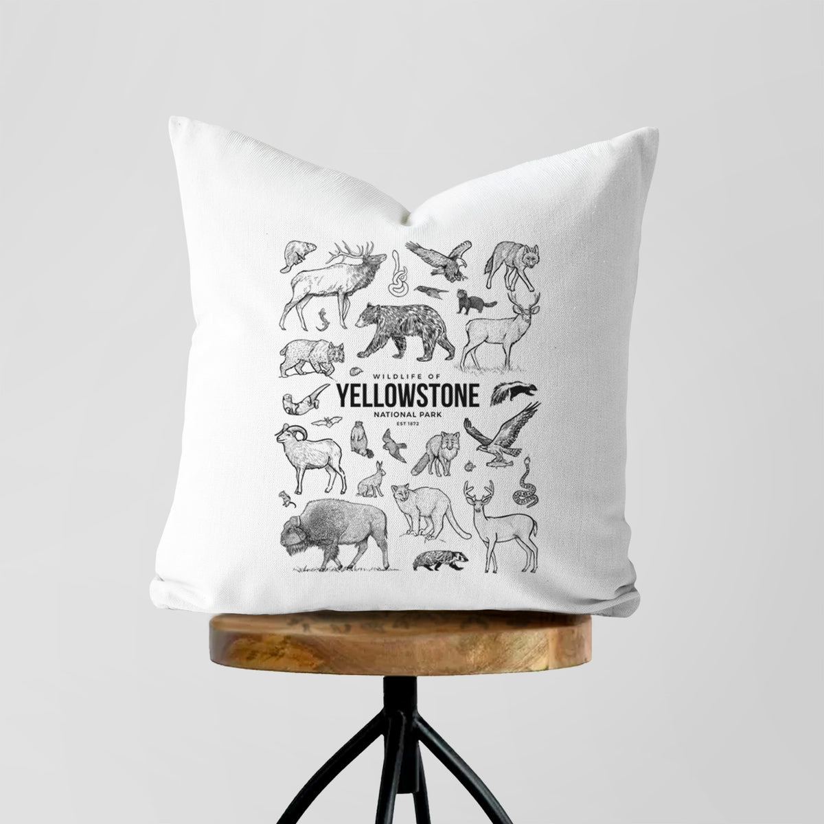 White pillow cover printed with a collection of black ink wildlife illustrations surrounding the words Yellowstone National Park, on a wooden stool.