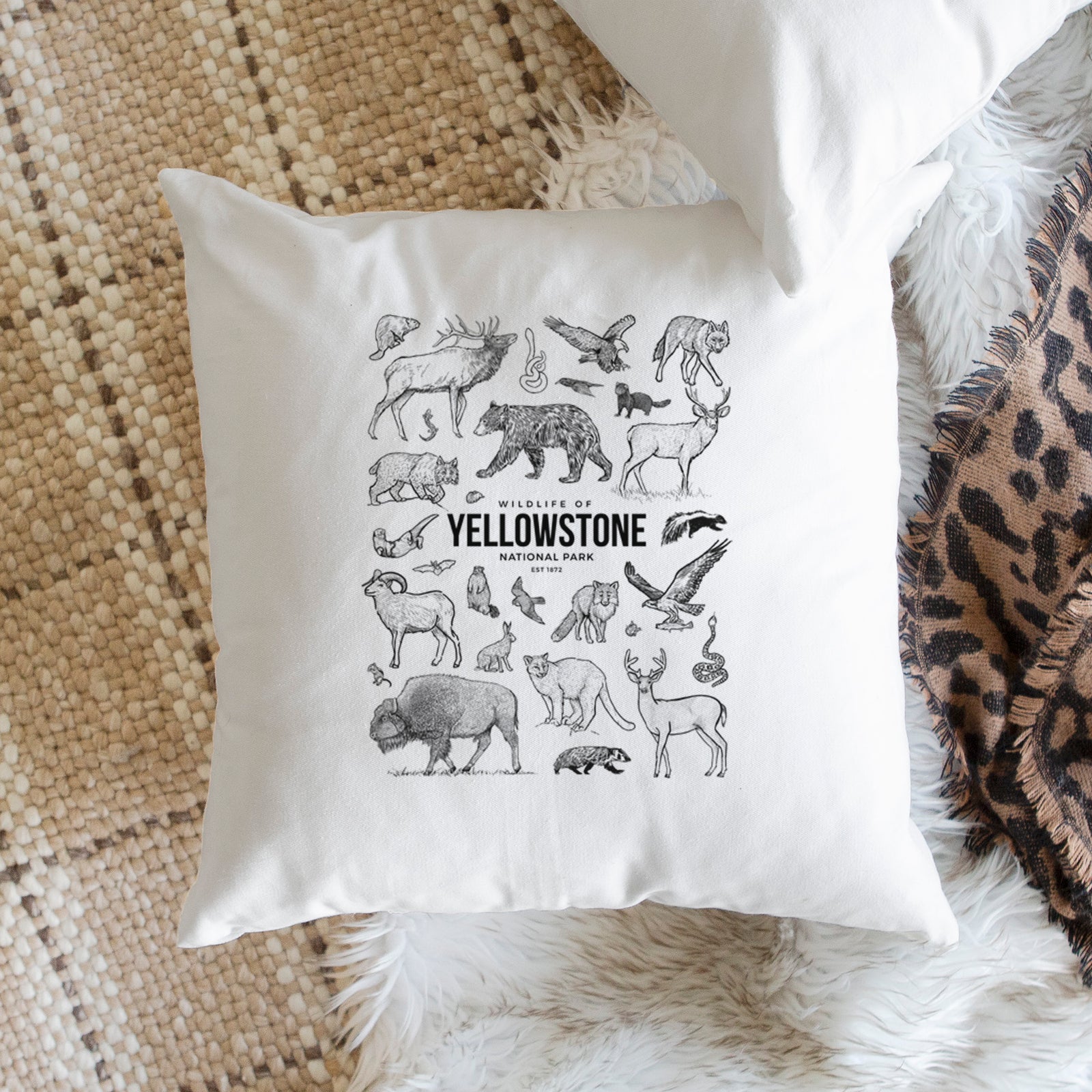 White pillow cover printed with a collection of black ink wildlife illustrations surrounding the words Yellowstone National Park, on a bench beside plants.