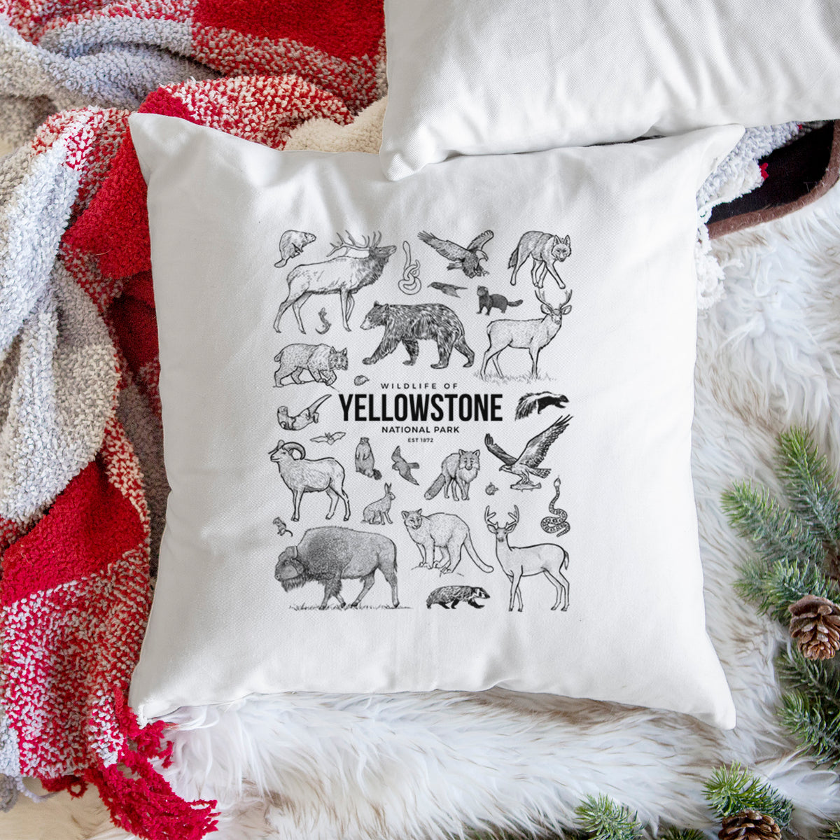 White pillow cover printed with a collection of black ink wildlife illustrations surrounding the words Yellowstone National Park, on a woven chair.