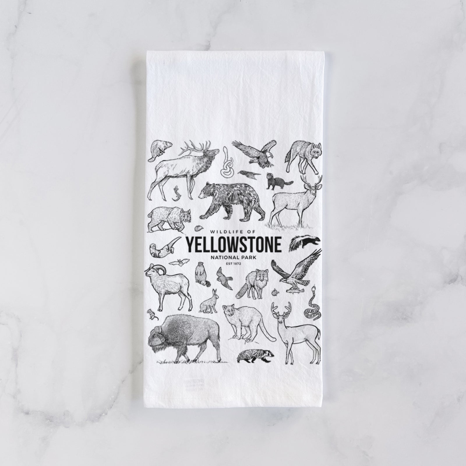 White cotton tea towel printed with a collection of black ink wildlife illustrations surrounding the words Yellowstone National Park, on a marble counter beside a round wooden cutting board and greenery.
