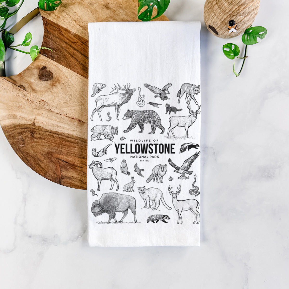 White cotton tea towel printed with a collection of black ink wildlife illustrations surrounding the words Yellowstone National Park, on a marble counter beside a round wooden cutting board and greenery.