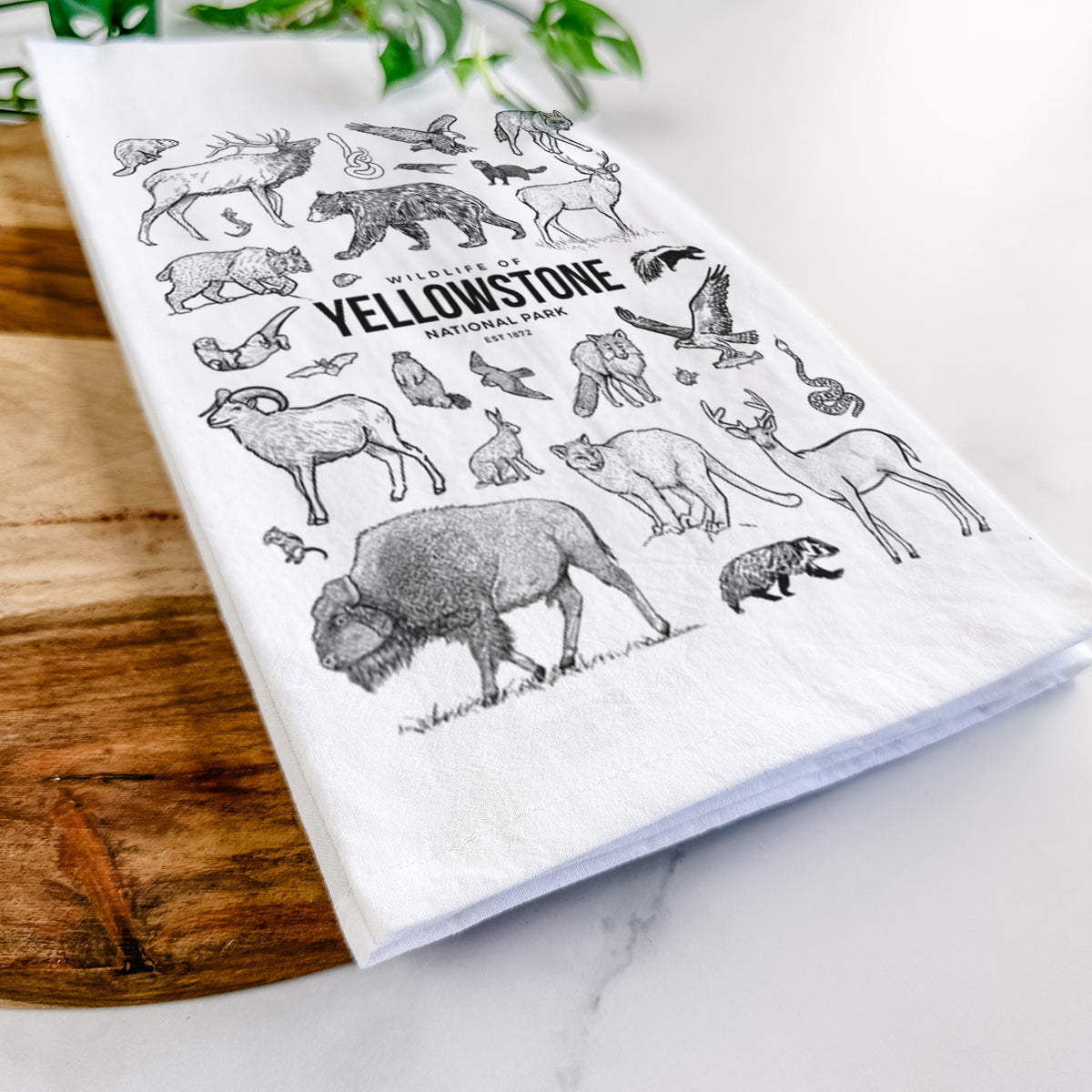 Close-up of a folded white tea towel on a wooden board, showing a collection of black ink wildlife illustrations surrounding the words Yellowstone National Park.