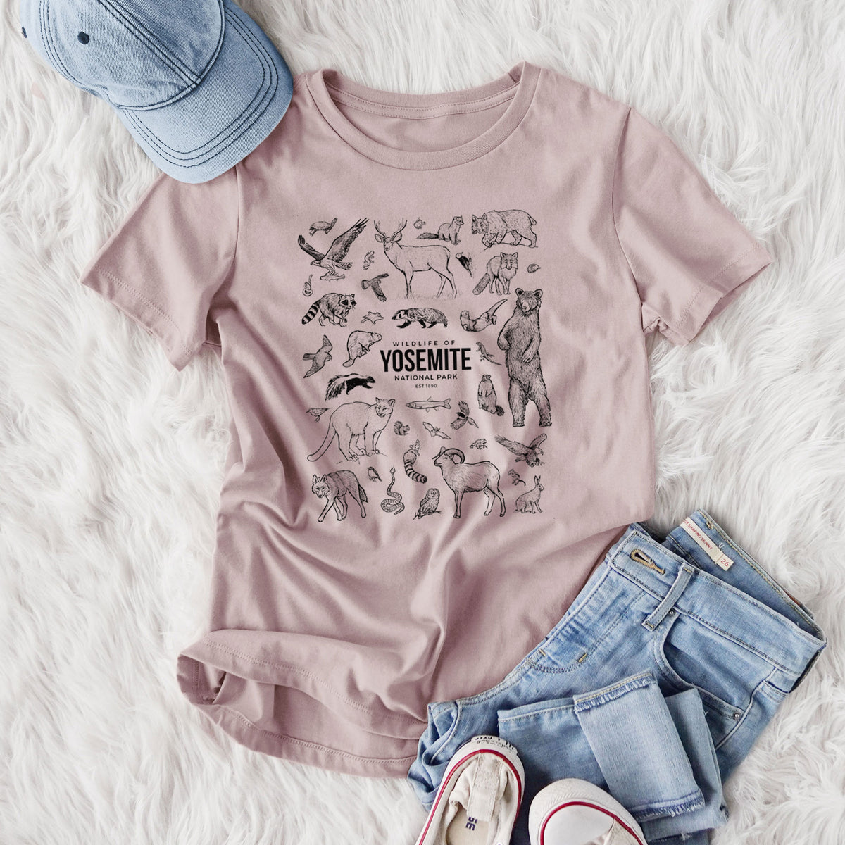 Women's relaxed fit cotton t-shirt printed with a collection of black ink wildlife illustrations surrounding the words Yosemite National Park, styled flat on a white faux fur rug with jeans.