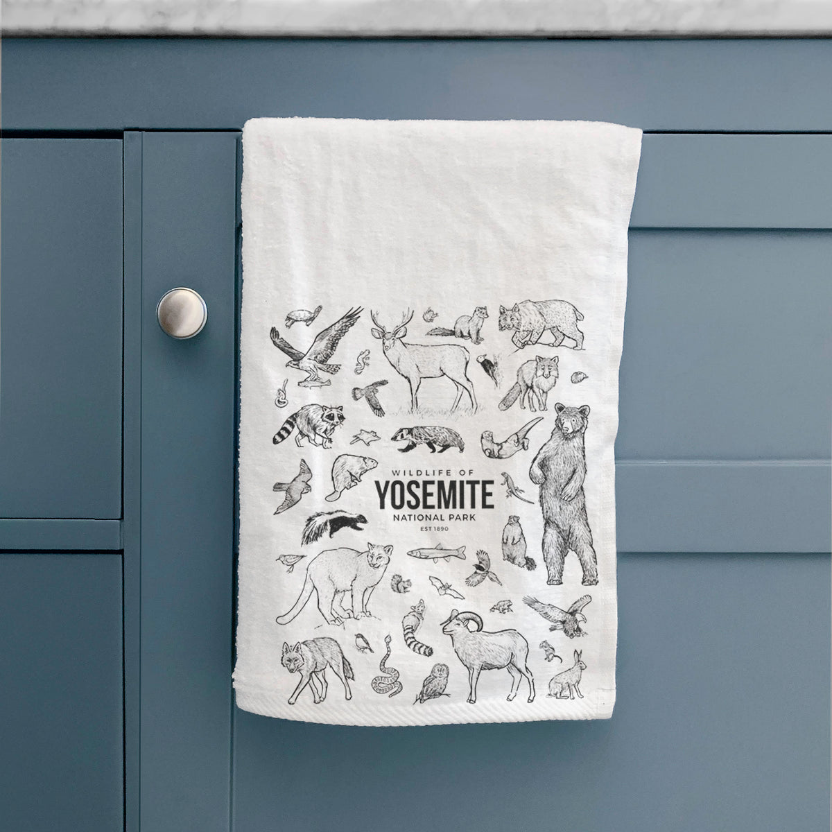White hand towel printed with a collection of black ink wildlife illustrations surrounding the words Yosemite National Park, hanging on a cabinet door.