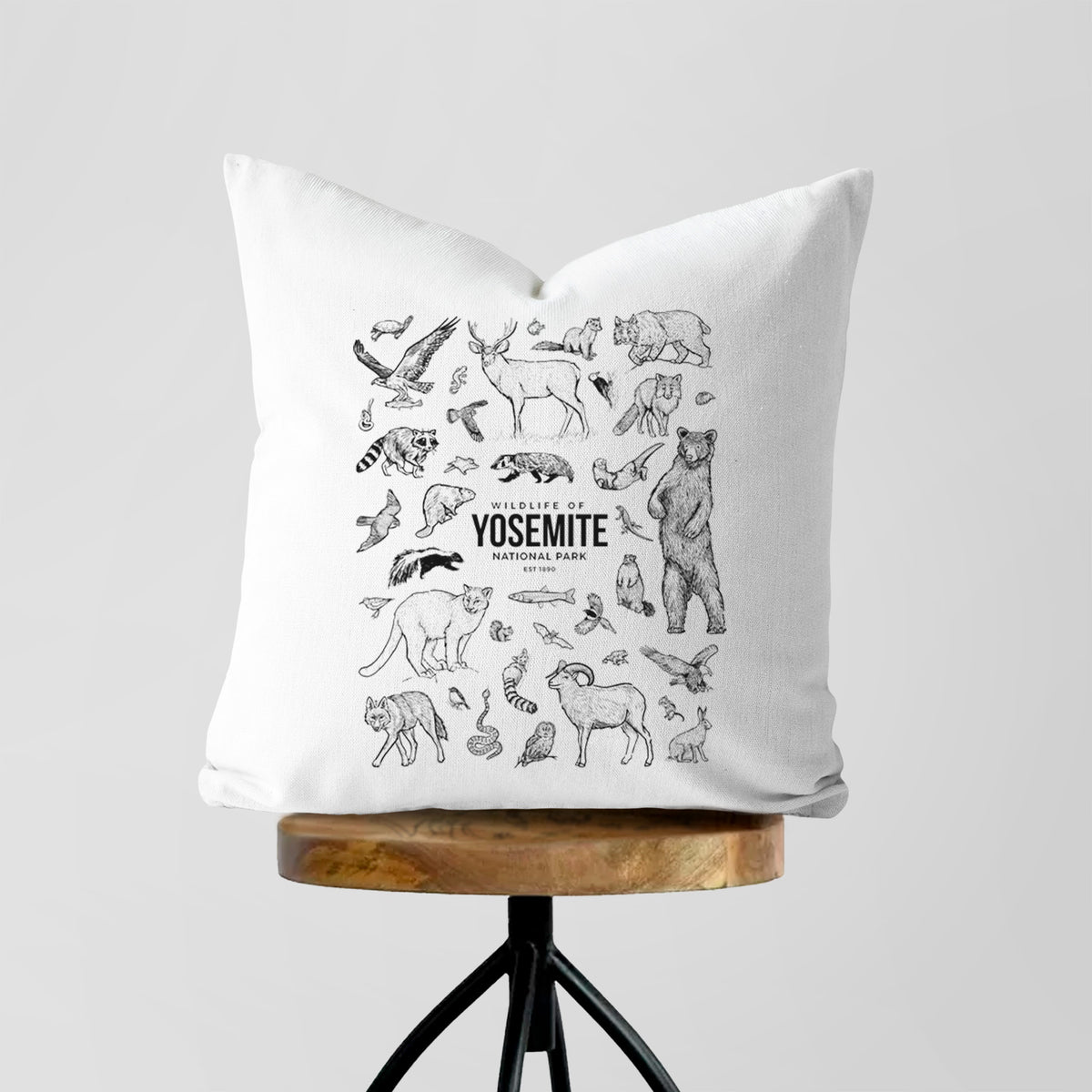 White pillow cover printed with a collection of black ink wildlife illustrations surrounding the words Yosemite National Park, on a wooden stool.