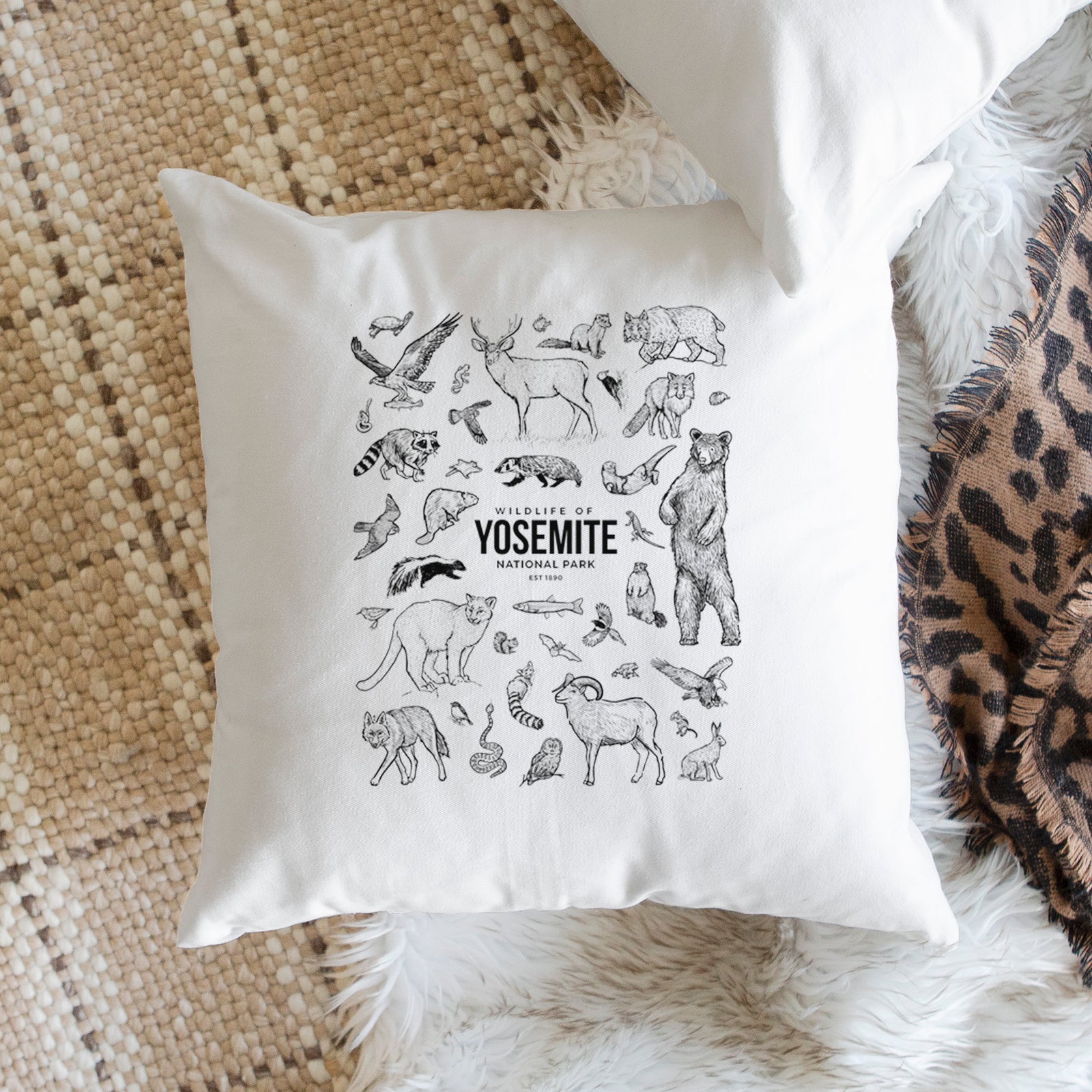 White pillow cover printed with a collection of black ink wildlife illustrations surrounding the words Yosemite National Park, on a bench beside plants.