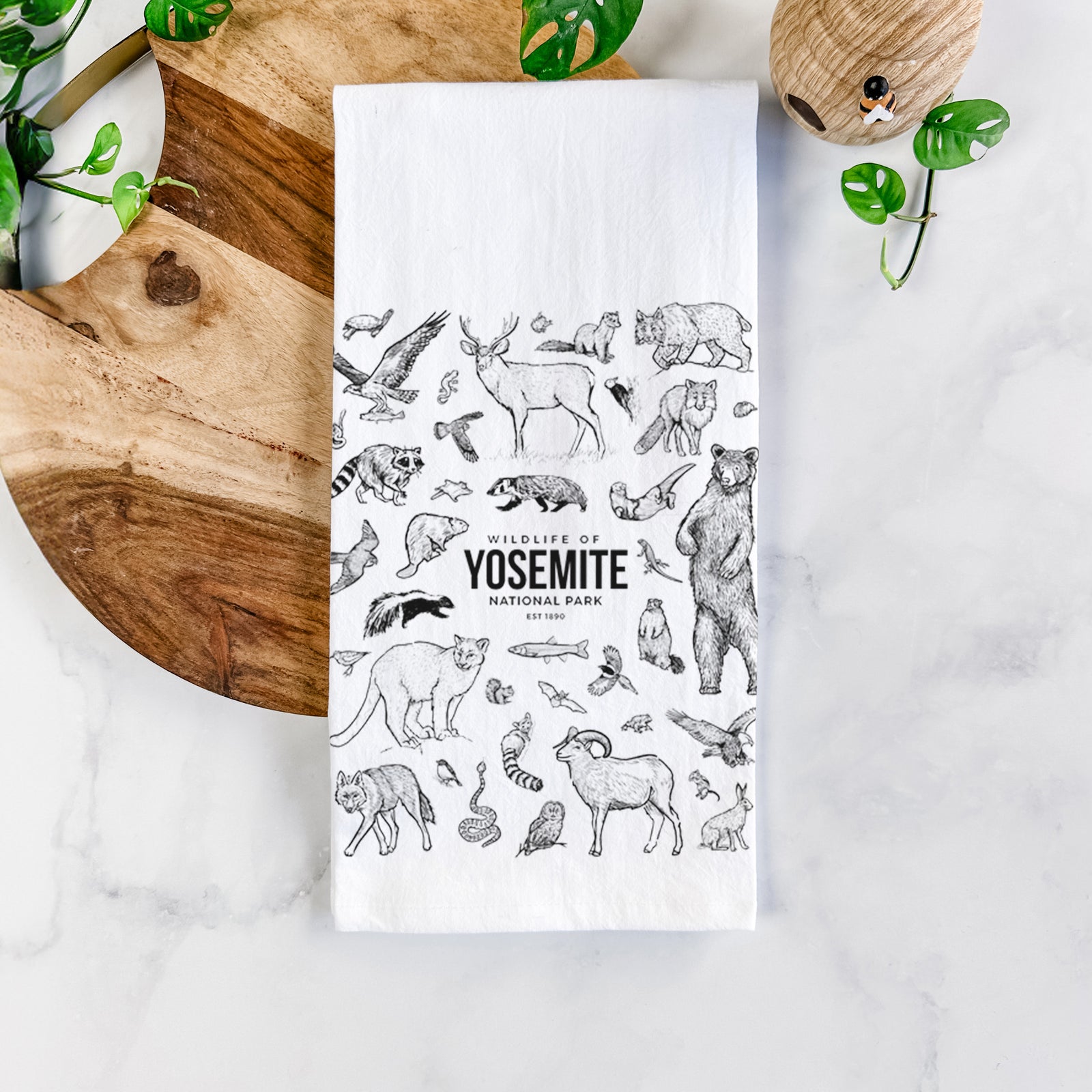 White cotton tea towel printed with a collection of black ink wildlife illustrations surrounding the words Yosemite National Park, on a marble counter beside a round wooden cutting board and greenery.