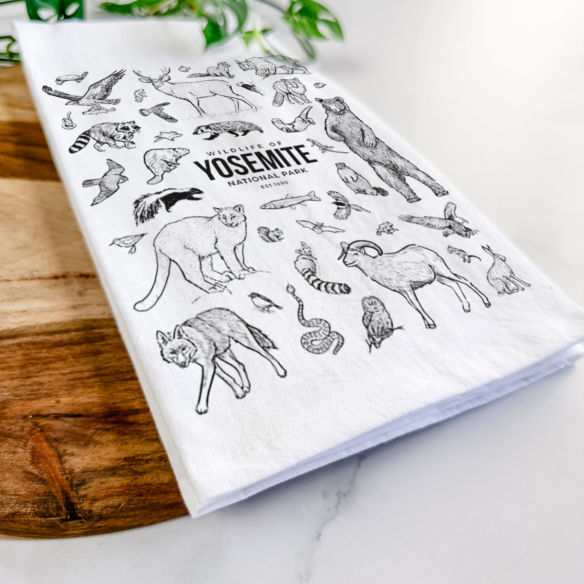 Close-up of a folded white tea towel on a wooden board, showing a collection of black ink wildlife illustrations surrounding the words Yosemite National Park.