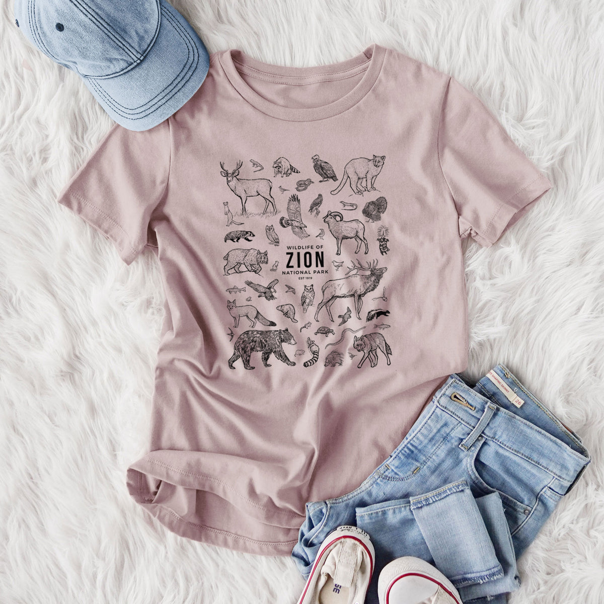 Women's relaxed fit cotton t-shirt printed with a collection of black ink wildlife illustrations surrounding the words Zion National Park, styled flat on a white faux fur rug with jeans.