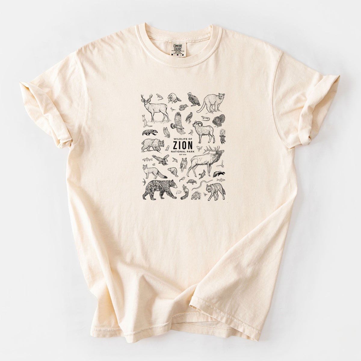 Ivory Comfort Colors t-shirt laid flat, printed with a collection of black ink wildlife illustrations surrounding the words Zion National Park.
