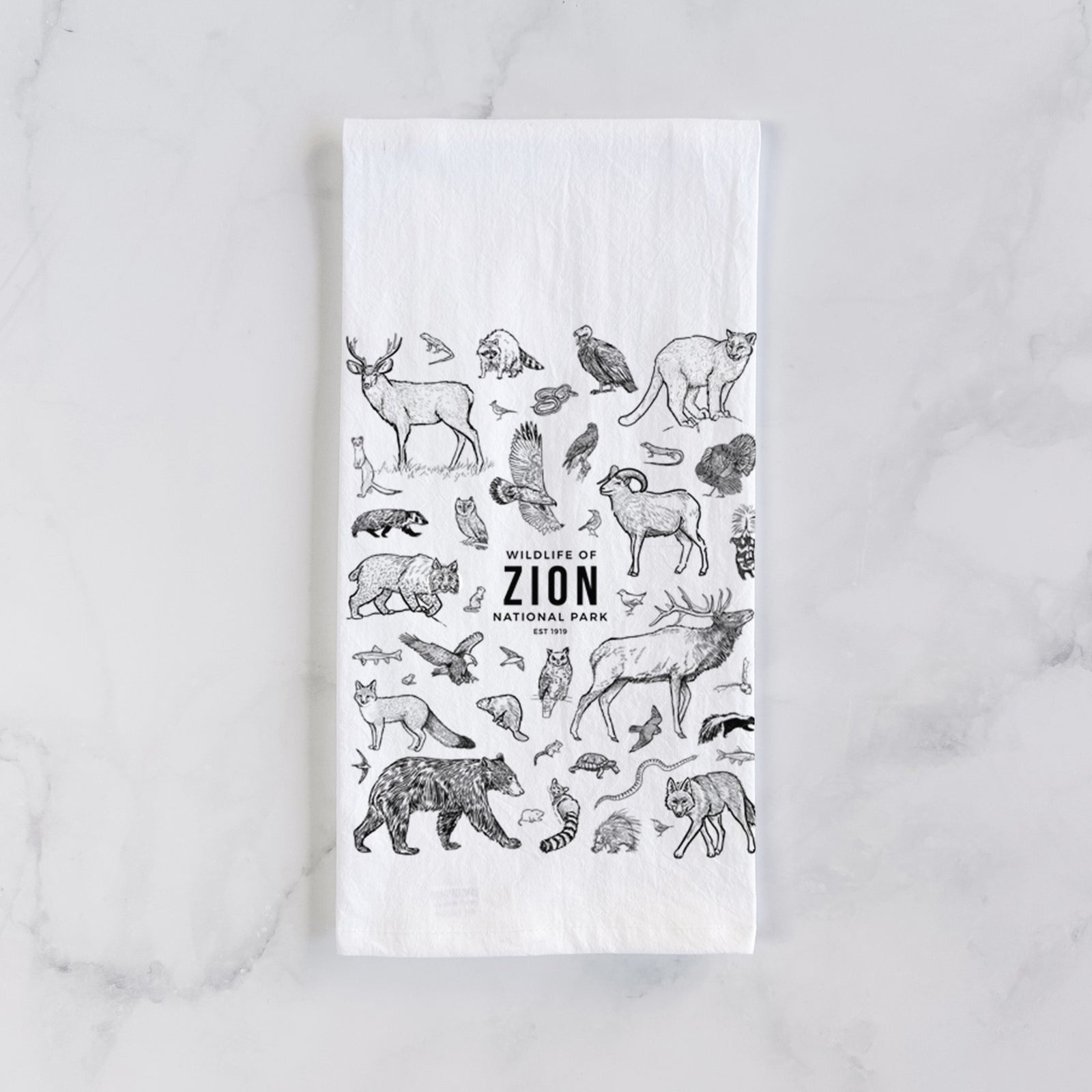 White cotton tea towel printed with a collection of black ink wildlife illustrations surrounding the words Zion National Park, on a marble counter beside a round wooden cutting board and greenery.