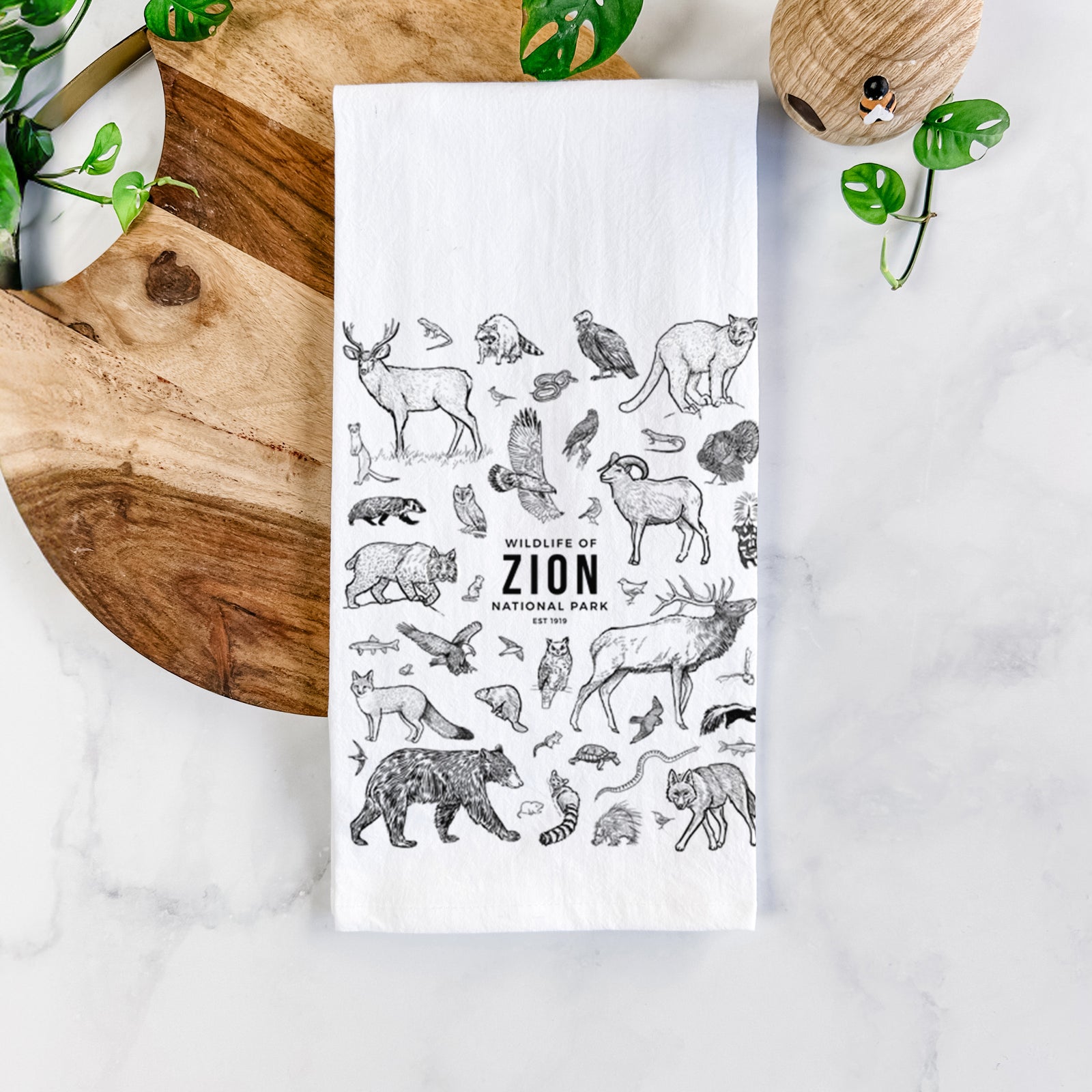 White cotton tea towel printed with a collection of black ink wildlife illustrations surrounding the words Zion National Park, on a marble counter beside a round wooden cutting board and greenery.
