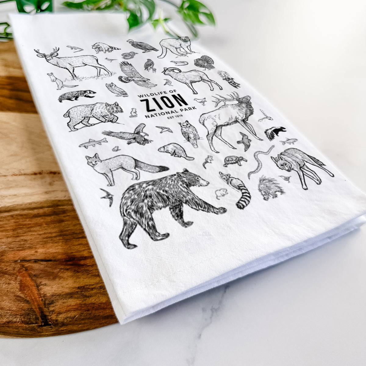 Close-up of a folded white tea towel on a wooden board, showing a collection of black ink wildlife illustrations surrounding the words Zion National Park.