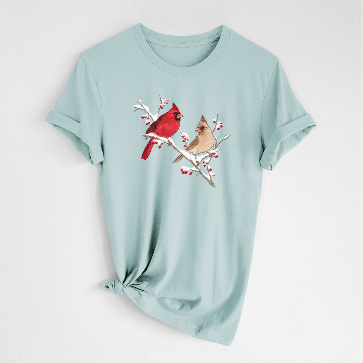 Dusty blue lightweight cotton t-shirt laid flat with the sleeves rolled, printed with a full-color illustration of a male and female northern cardinal perched on a snowy branch with red berries.