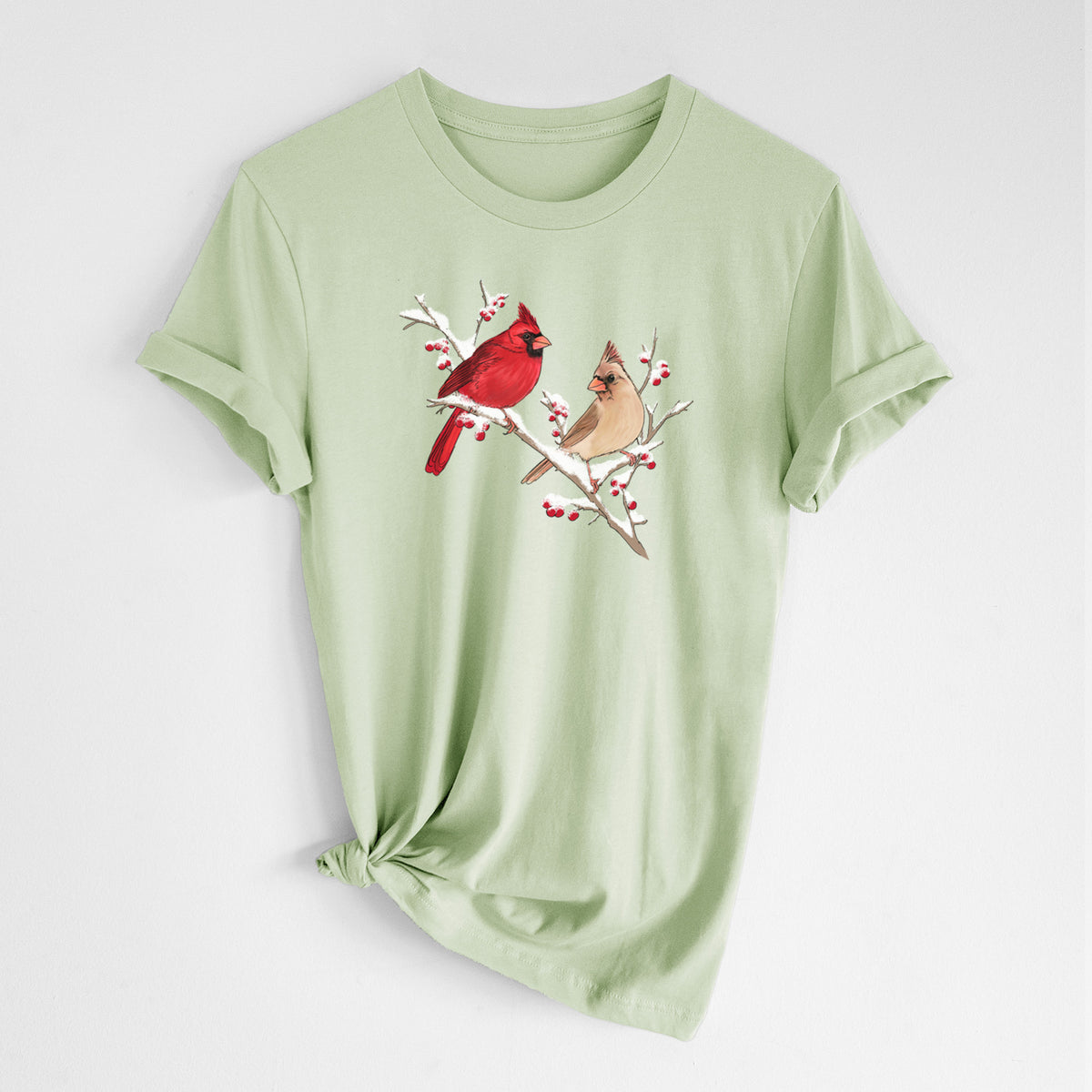 Spring green lightweight cotton t-shirt laid flat with the sleeves rolled, printed with a full-color illustration of a male and female northern cardinal perched on a snowy branch with red berries.