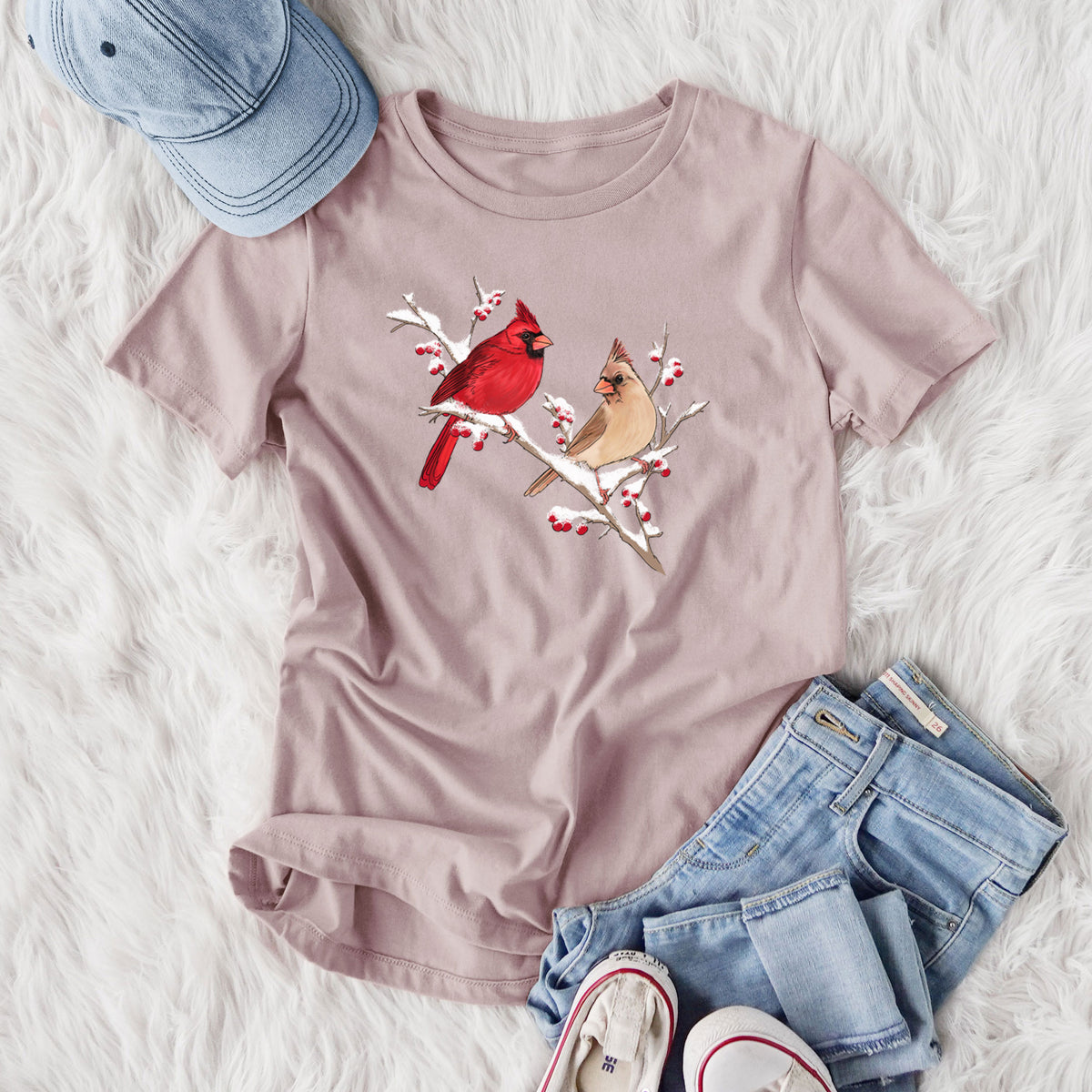 Women's relaxed fit cotton t-shirt printed with a full-color illustration of a male and female northern cardinal perched on a snowy branch with red berries, styled flat on a white faux fur rug with jeans.