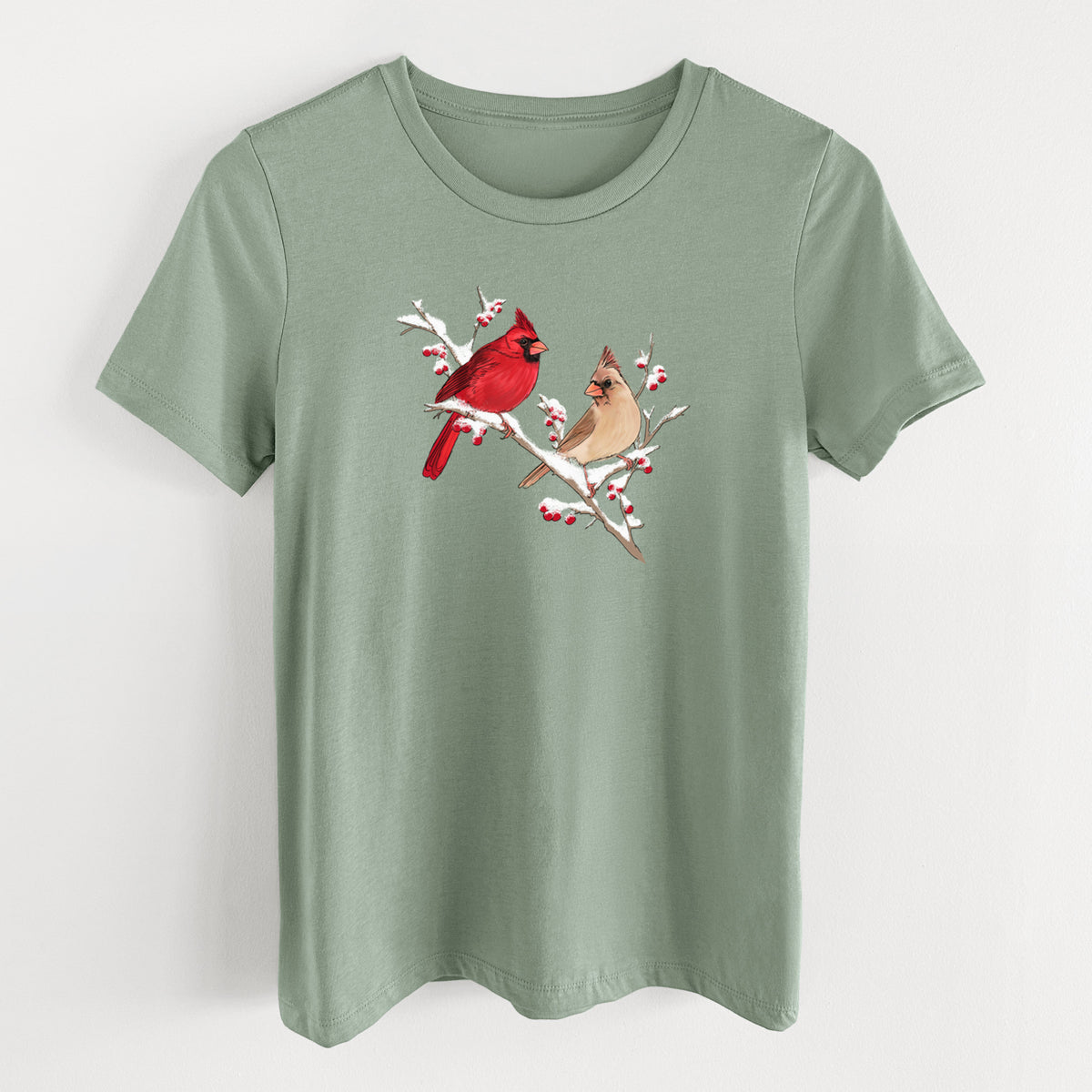Sage green women's relaxed fit cotton t-shirt laid flat, printed with a full-color illustration of a male and female northern cardinal perched on a snowy branch with red berries.