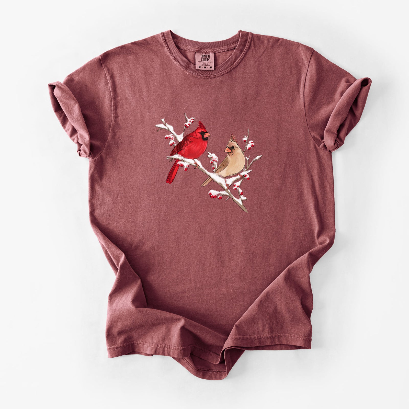 Brick red Comfort Colors t-shirt laid flat, printed with a full-color illustration of a male and female northern cardinal perched on a snowy branch with red berries.