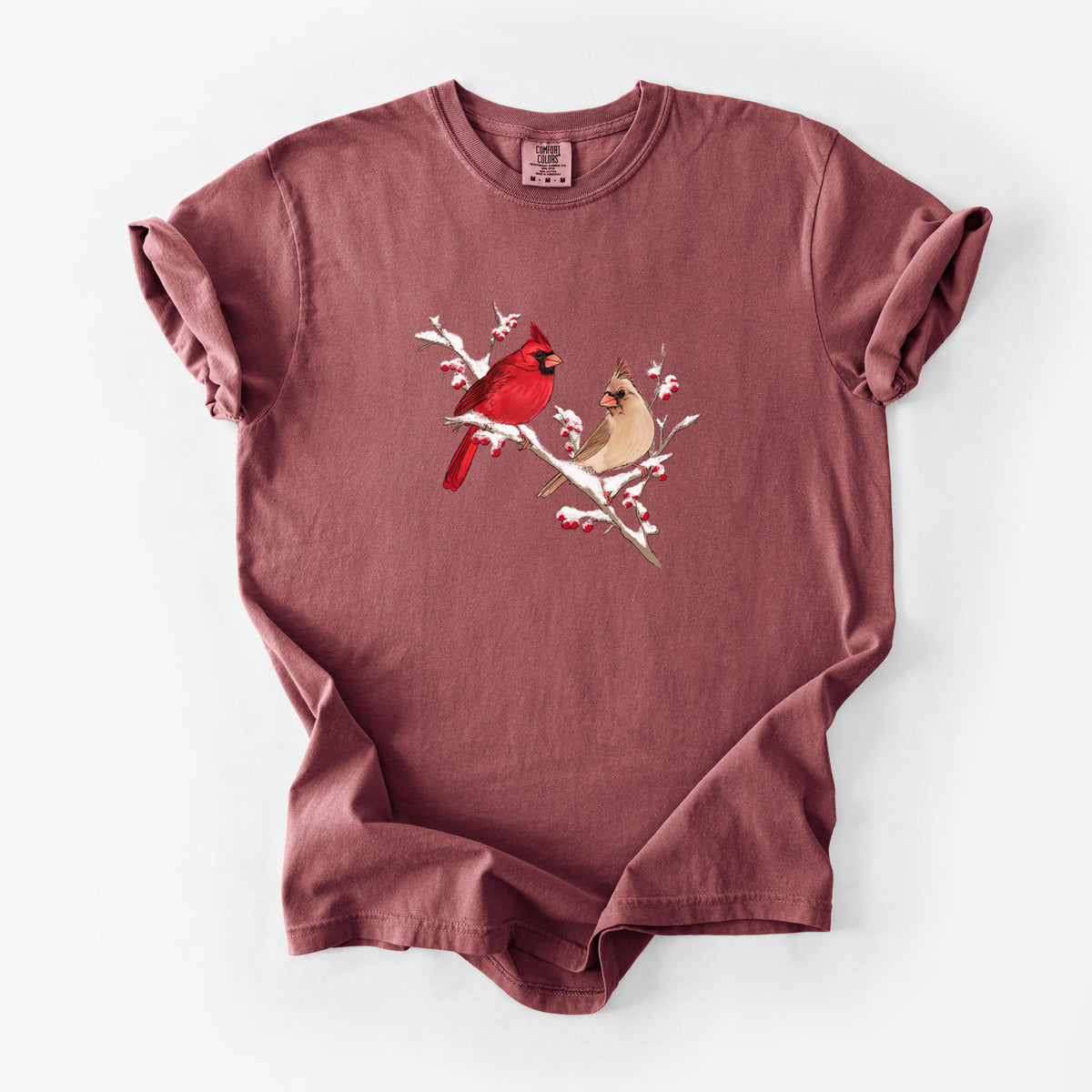 Brick red Comfort Colors t-shirt laid flat, printed with a full-color illustration of a male and female northern cardinal perched on a snowy branch with red berries.