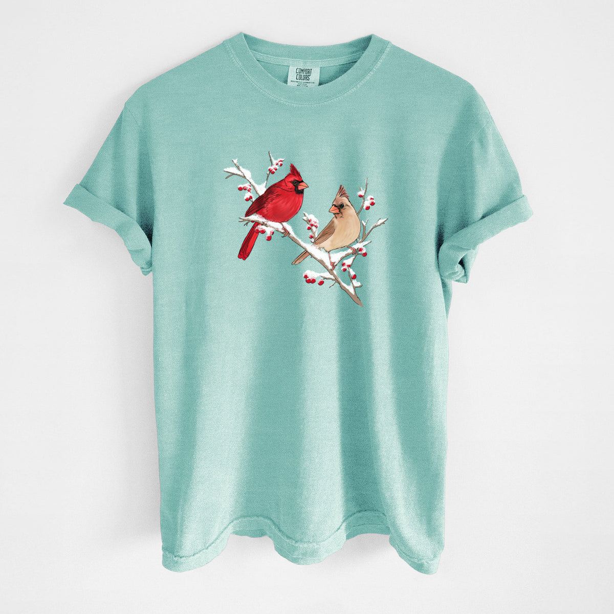 Chalky mint Comfort Colors t-shirt laid flat, printed with a full-color illustration of a male and female northern cardinal perched on a snowy branch with red berries.