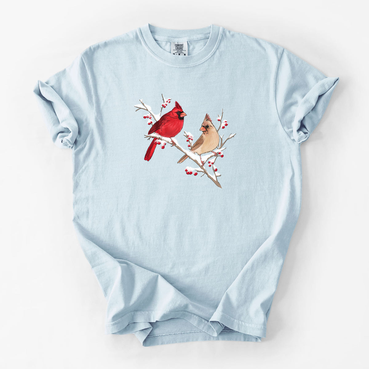 Chambray blue Comfort Colors t-shirt laid flat, printed with a full-color illustration of a male and female northern cardinal perched on a snowy branch with red berries.