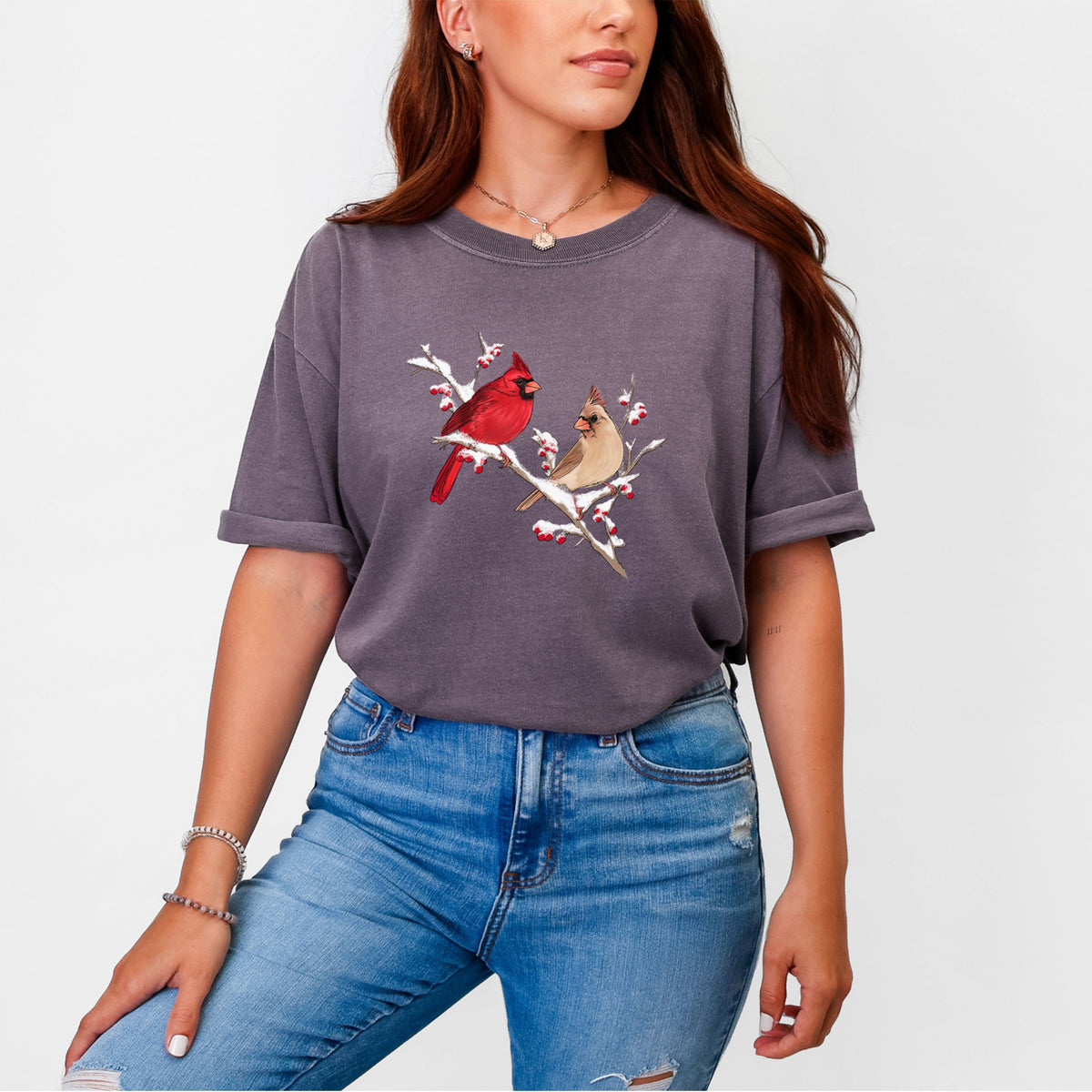 Woman wearing a Comfort Colors t-shirt printed with a full-color illustration of a male and female northern cardinal perched on a snowy branch with red berries.