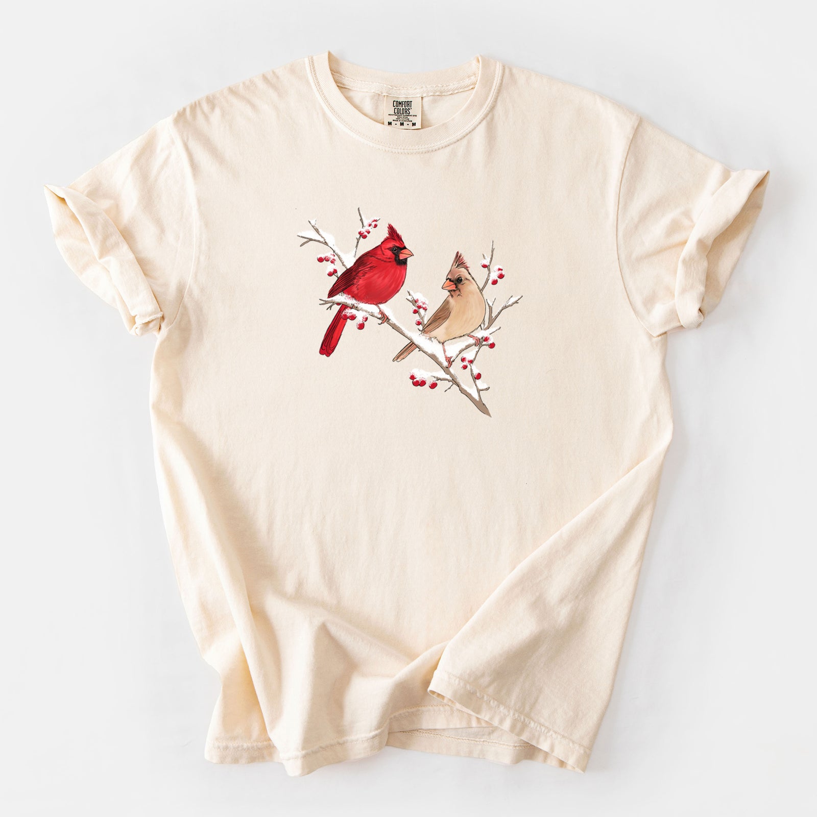 Brick red Comfort Colors t-shirt laid flat, printed with a full-color illustration of a male and female northern cardinal perched on a snowy branch with red berries.