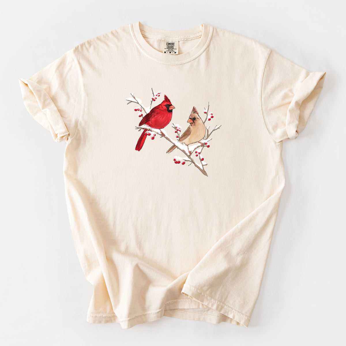 Ivory Comfort Colors t-shirt laid flat, printed with a full-color illustration of a male and female northern cardinal perched on a snowy branch with red berries.