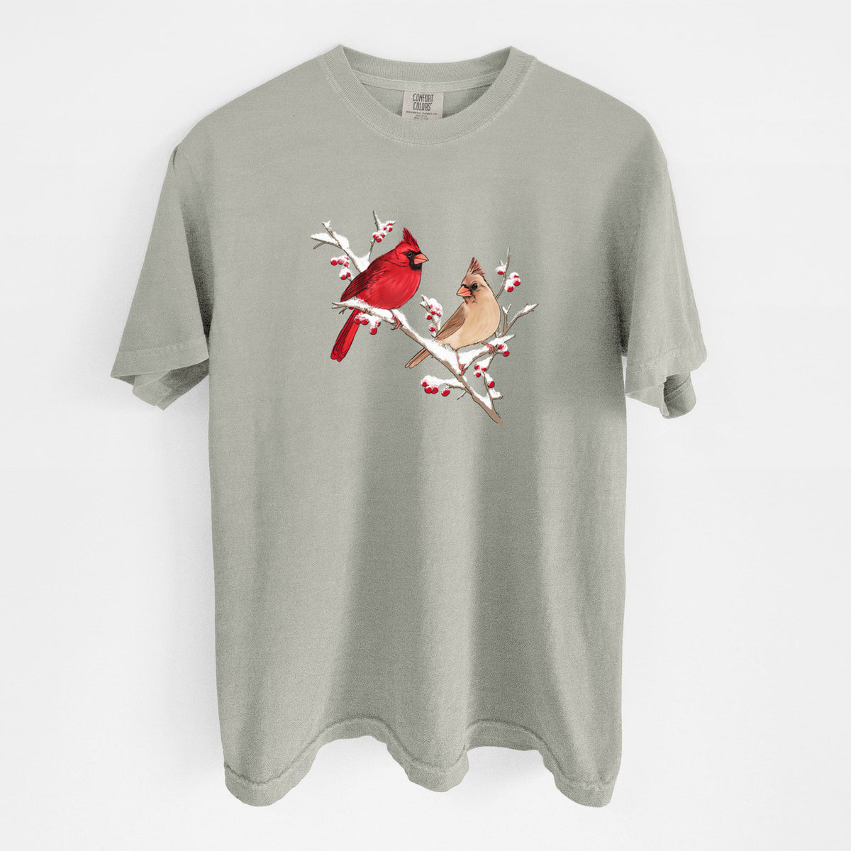Sandstone beige Comfort Colors t-shirt laid flat, printed with a full-color illustration of a male and female northern cardinal perched on a snowy branch with red berries.