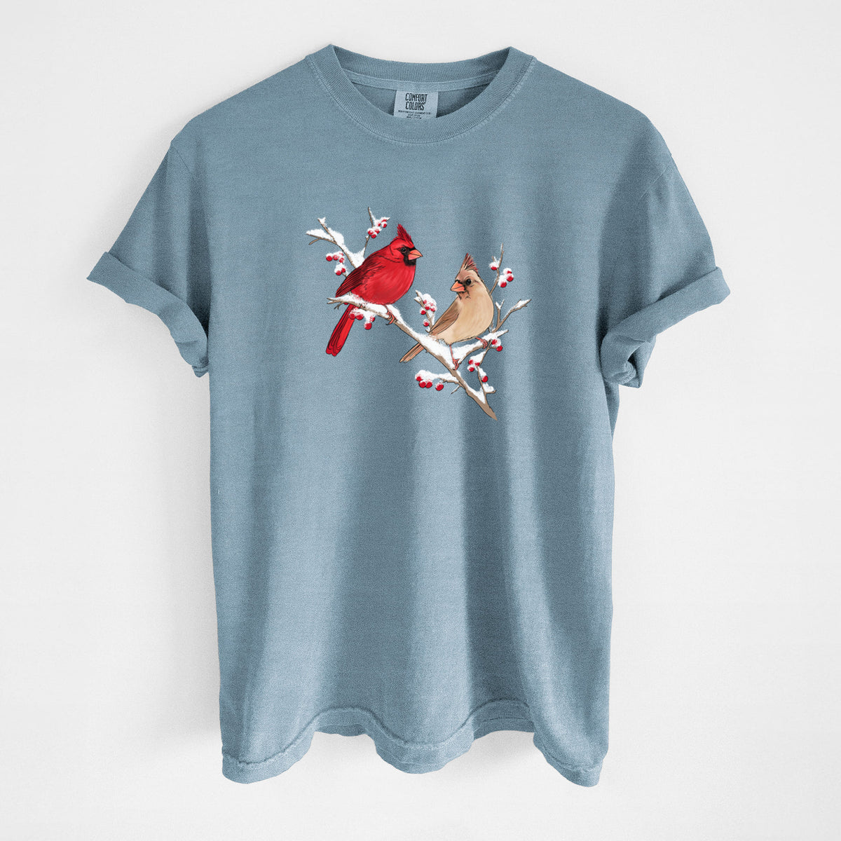 Steel blue Comfort Colors t-shirt laid flat, printed with a full-color illustration of a male and female northern cardinal perched on a snowy branch with red berries.