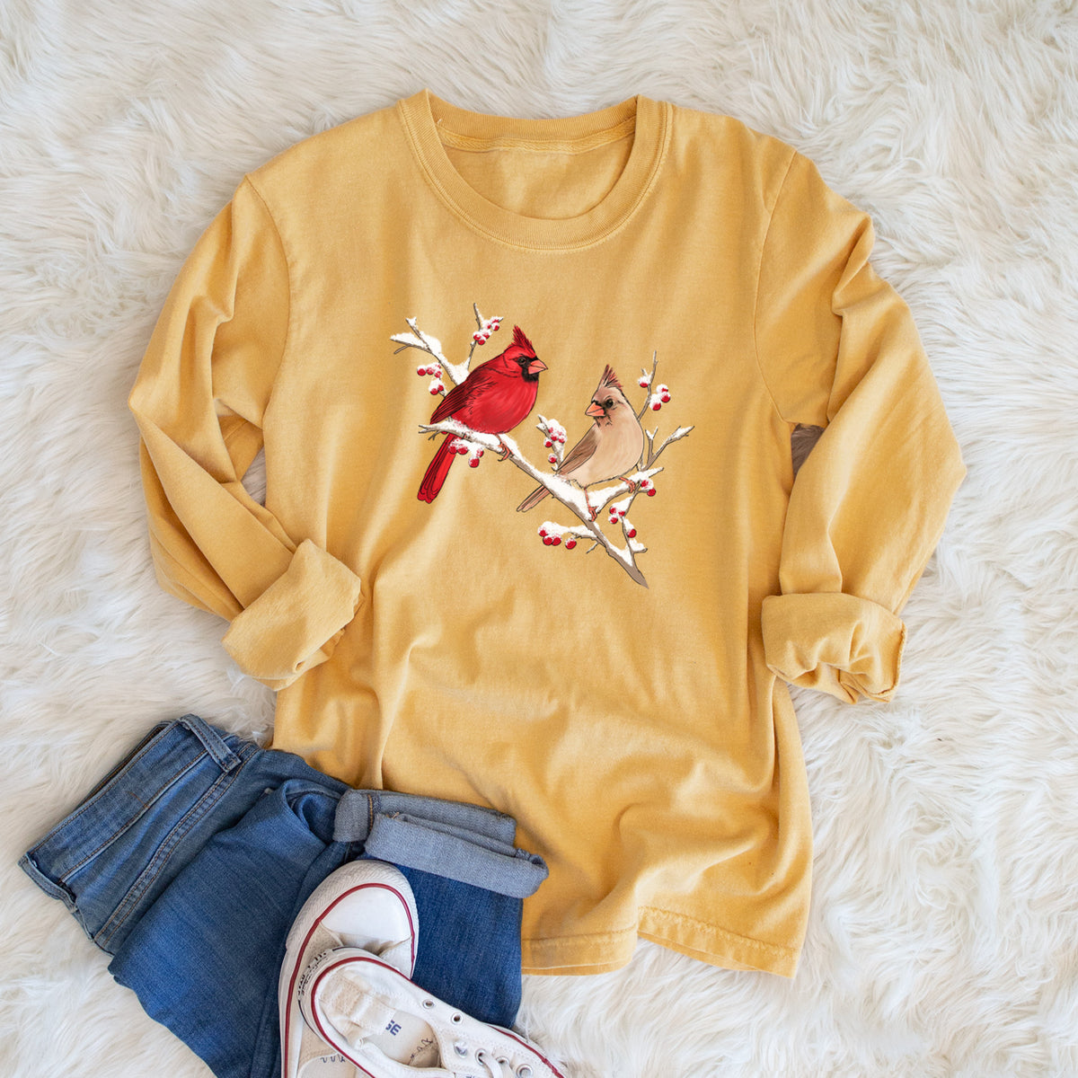Golden wheat yellow Comfort Colors long sleeve shirt printed with a full-color illustration of a male and female northern cardinal perched on a snowy branch with red berries, shown in a lifestyle photo.