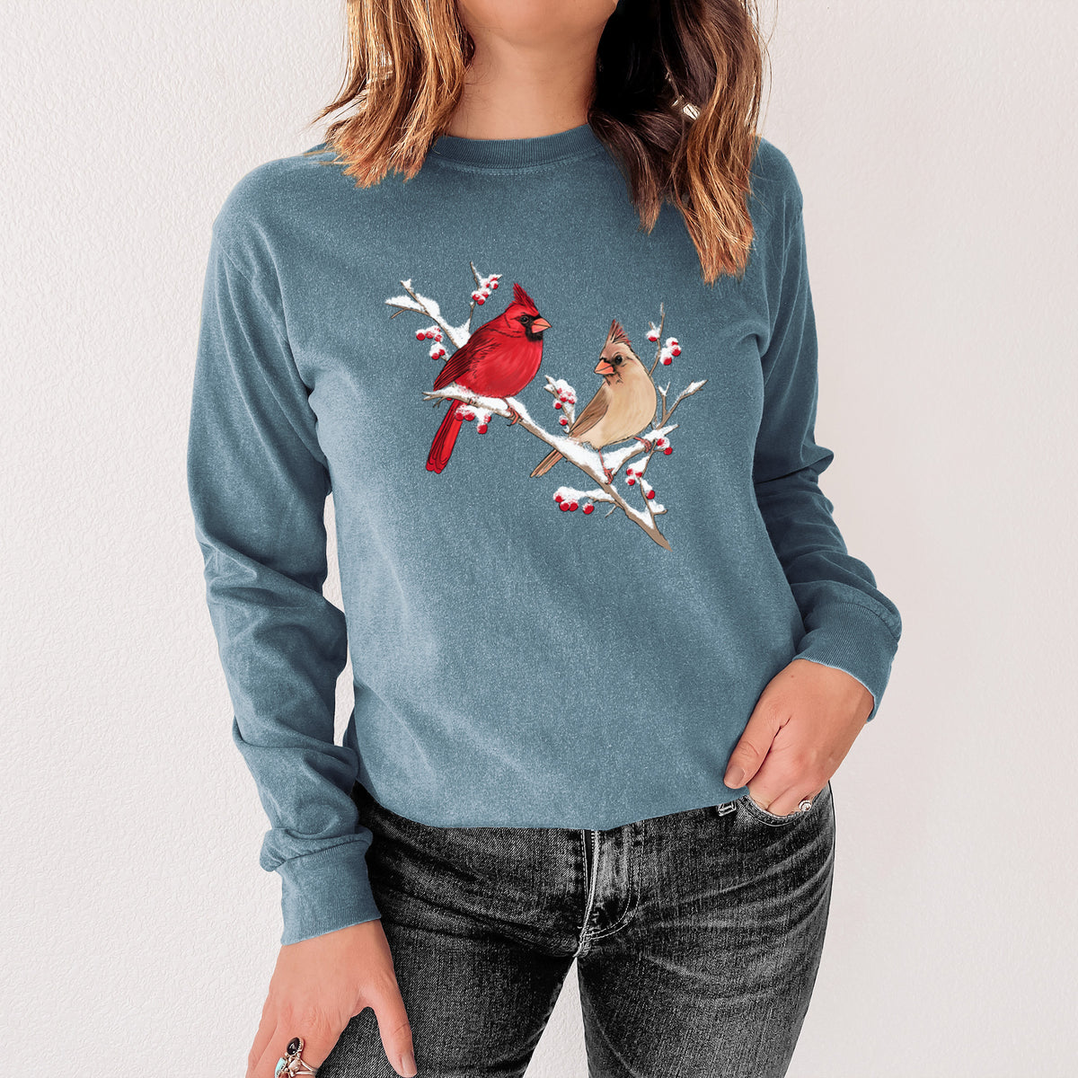Steel blue Comfort Colors long sleeve shirt printed with a full-color illustration of a male and female northern cardinal perched on a snowy branch with red berries, shown in a lifestyle photo.