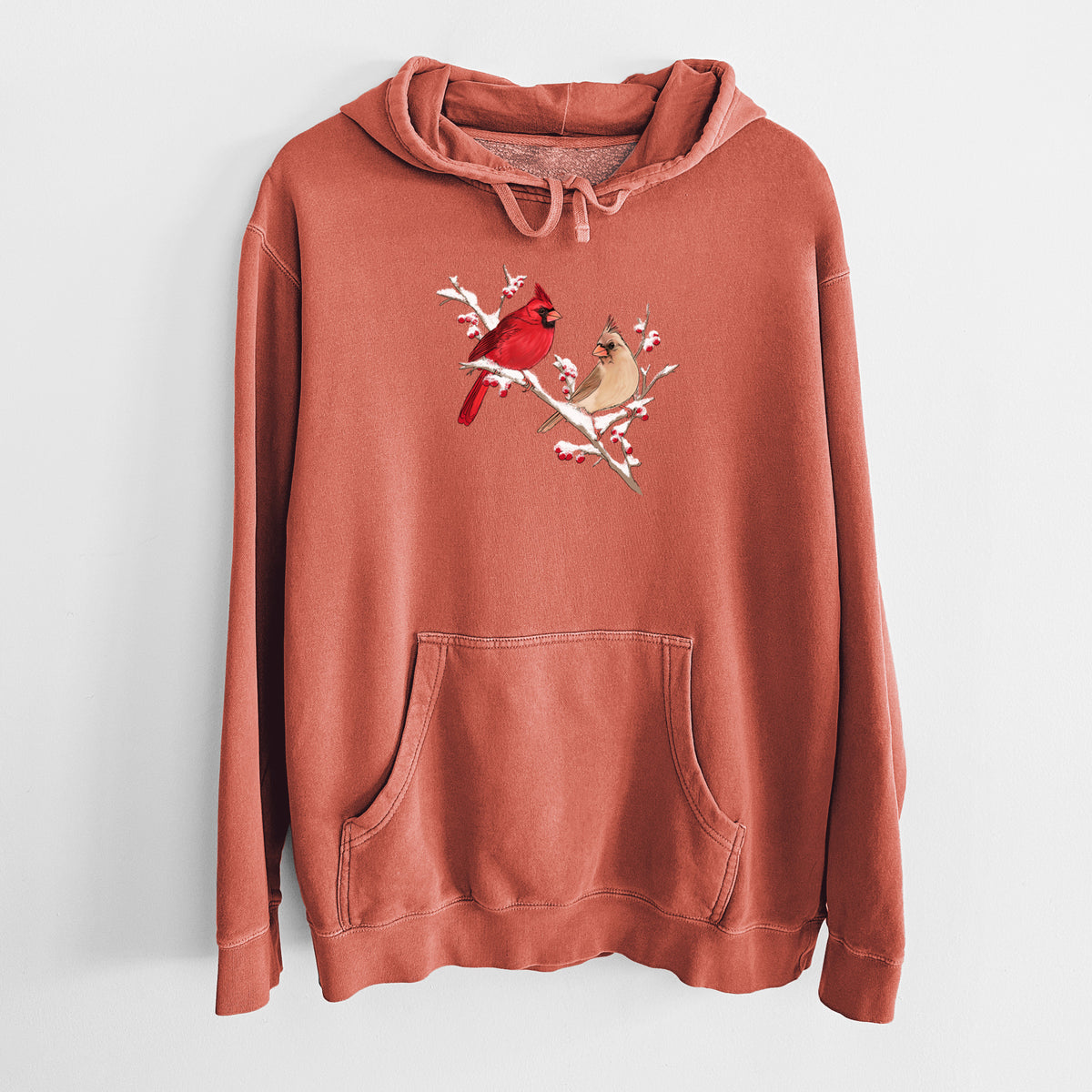 Amber orange pigment dyed hoodie laid flat, printed with a full-color illustration of a male and female northern cardinal perched on a snowy branch with red berries.