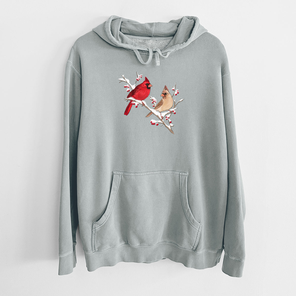 Sage green pigment dyed hoodie laid flat, printed with a full-color illustration of a male and female northern cardinal perched on a snowy branch with red berries.